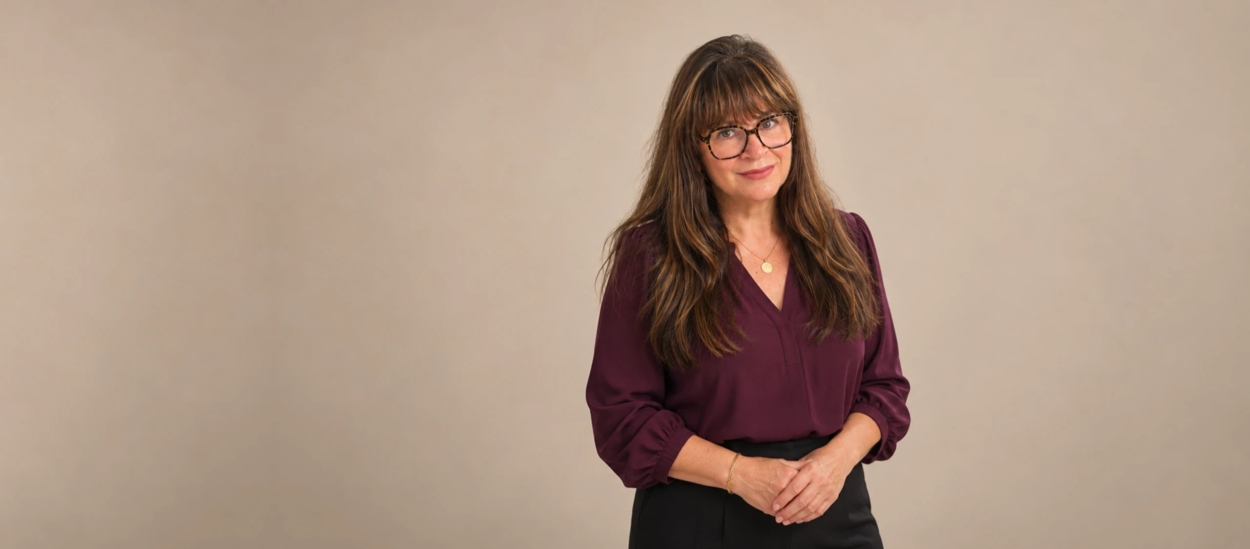 Dr. Alison Feit standing against a warm beige backdrop, wearing a plum blouse and tortoiseshell glasses